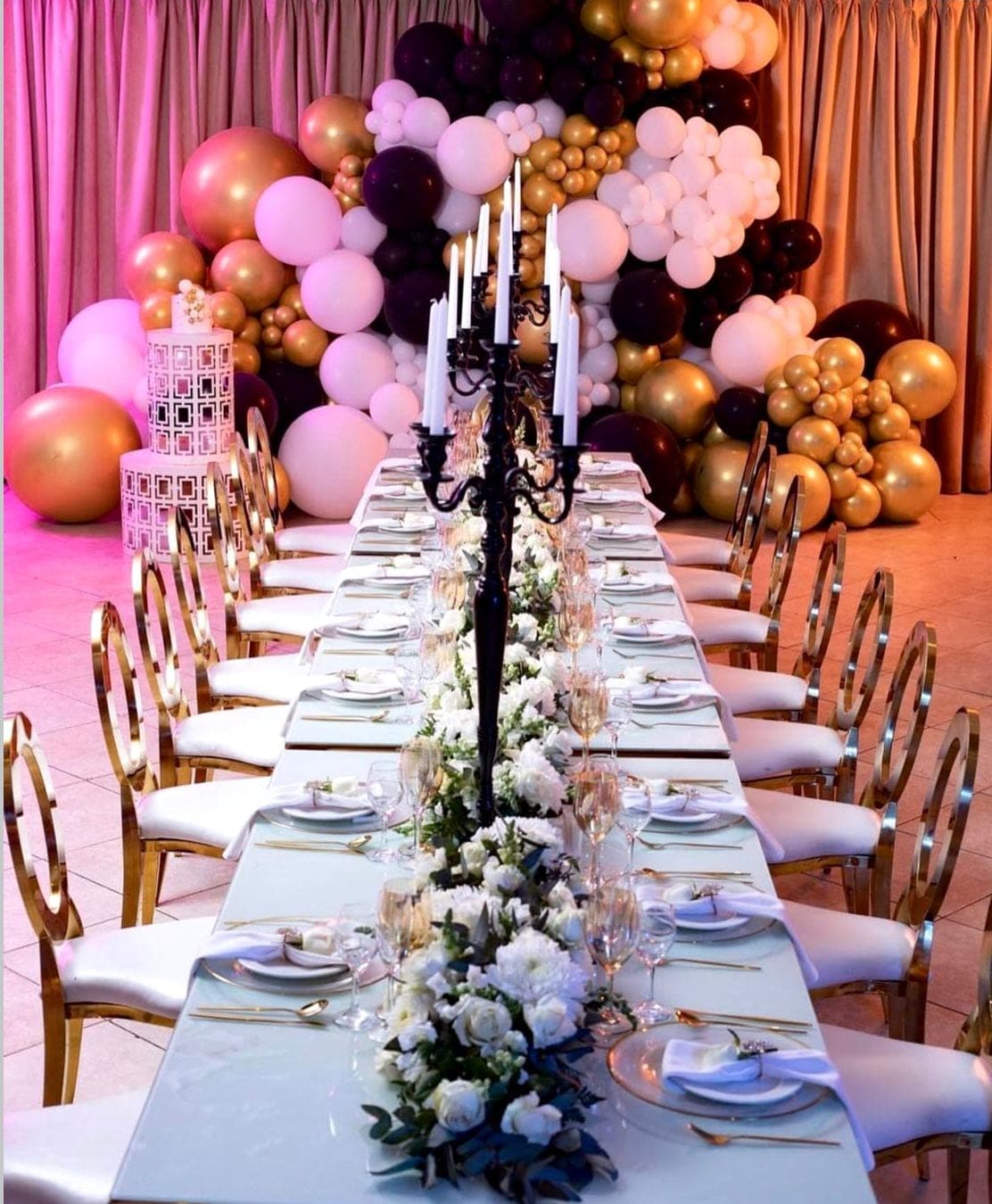 Long candle-lit tablescape with black candelabra, gold and black balloon arch, and gold oval-back chairs by Frontrow Creatives